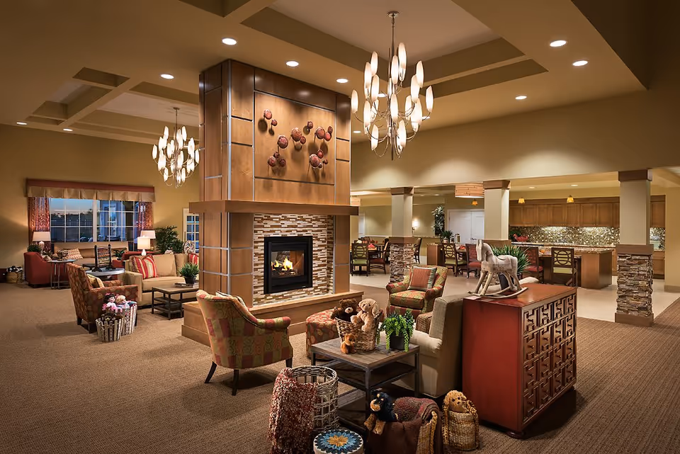 Spacious, warmly lit communal living room with seating arranged around a central fireplace and an adjacent dining/kitchen area.