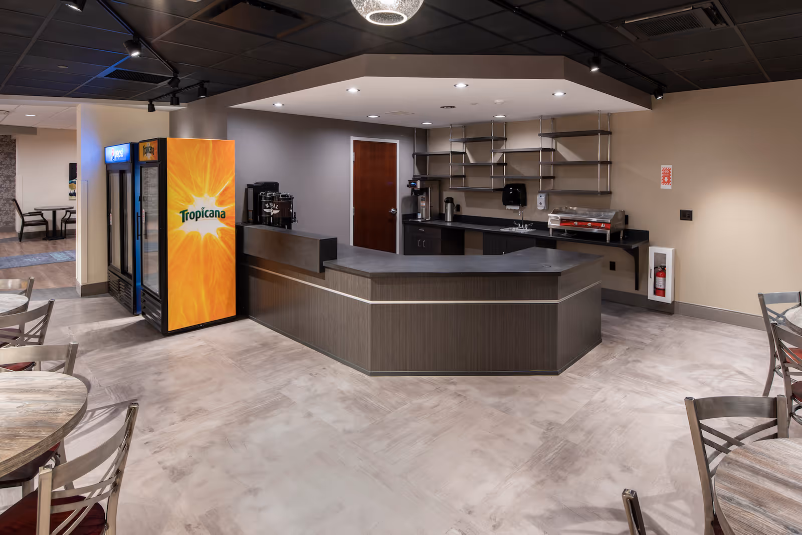 Modern cafeteria-style serving counter with a Tropicana beverage cooler and round dining tables in a communal dining area.