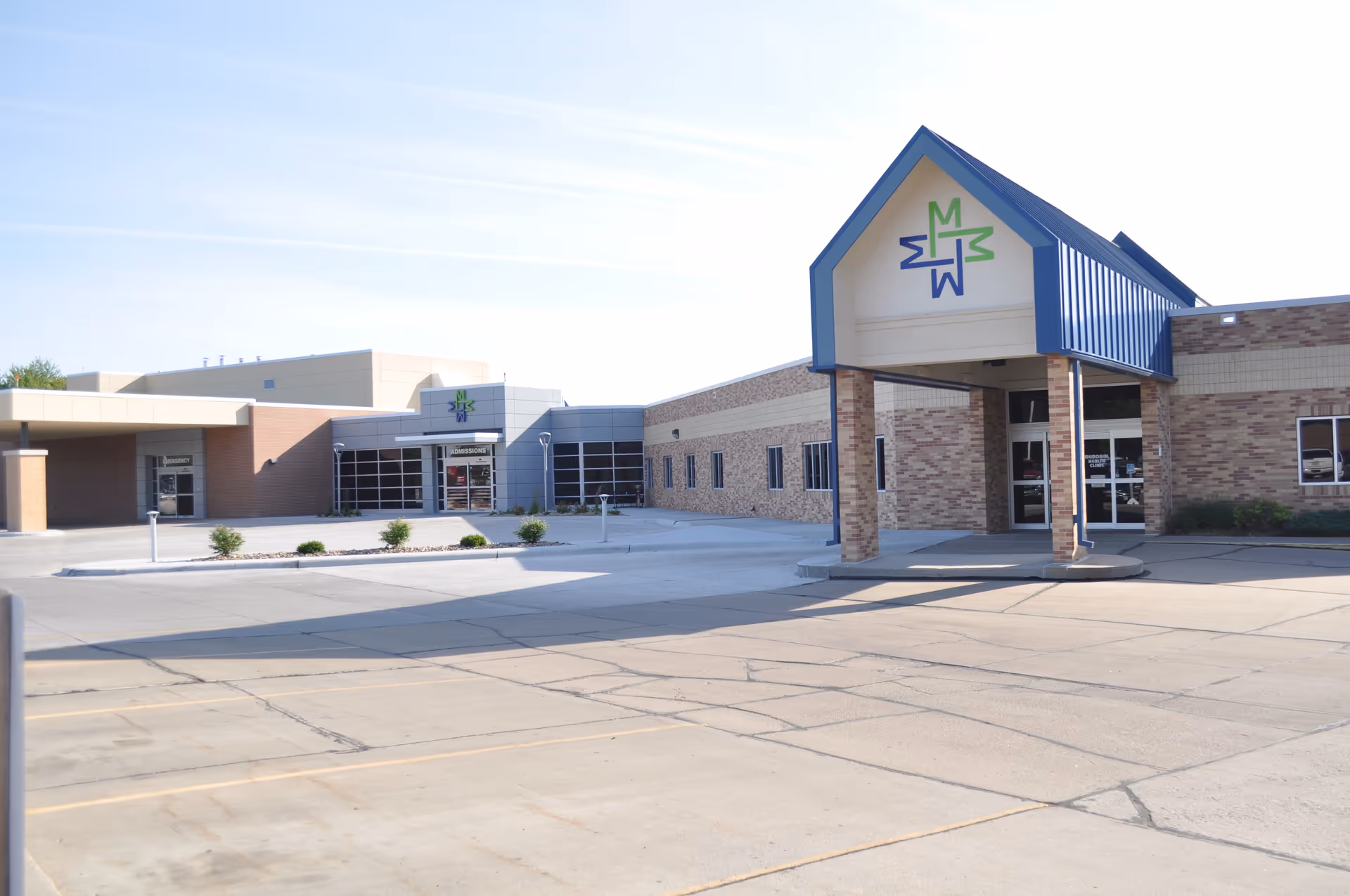 Exterior view of Memorial Community Health building with a covered entrance featuring a blue roof and a logo with green and blue letters. The building has a brick and beige facade with multiple windows and a large parking area in front.