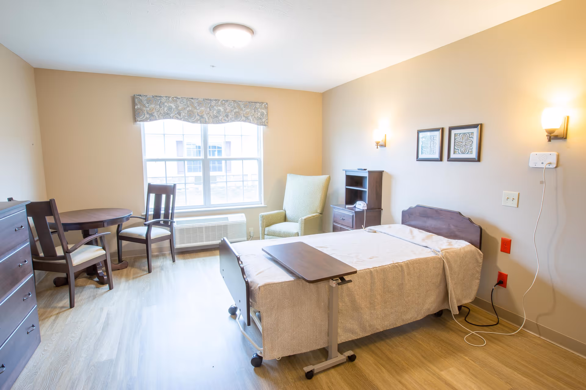 A well-lit bedroom in a senior living facility featuring a single bed with a beige blanket and an adjustable overbed table. The room includes a green upholstered armchair, a small wooden table with two chairs near a window, a wooden dresser, and two framed pictures on the wall. The floor is light wood, and the walls are painted a soft beige color.
