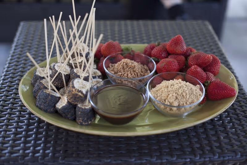A green plate on a woven table holding several skewered marshmallows coated in chocolate and crushed cookies, fresh strawberries, and three small glass bowls containing chocolate sauce, crushed nuts, and crushed graham crackers.