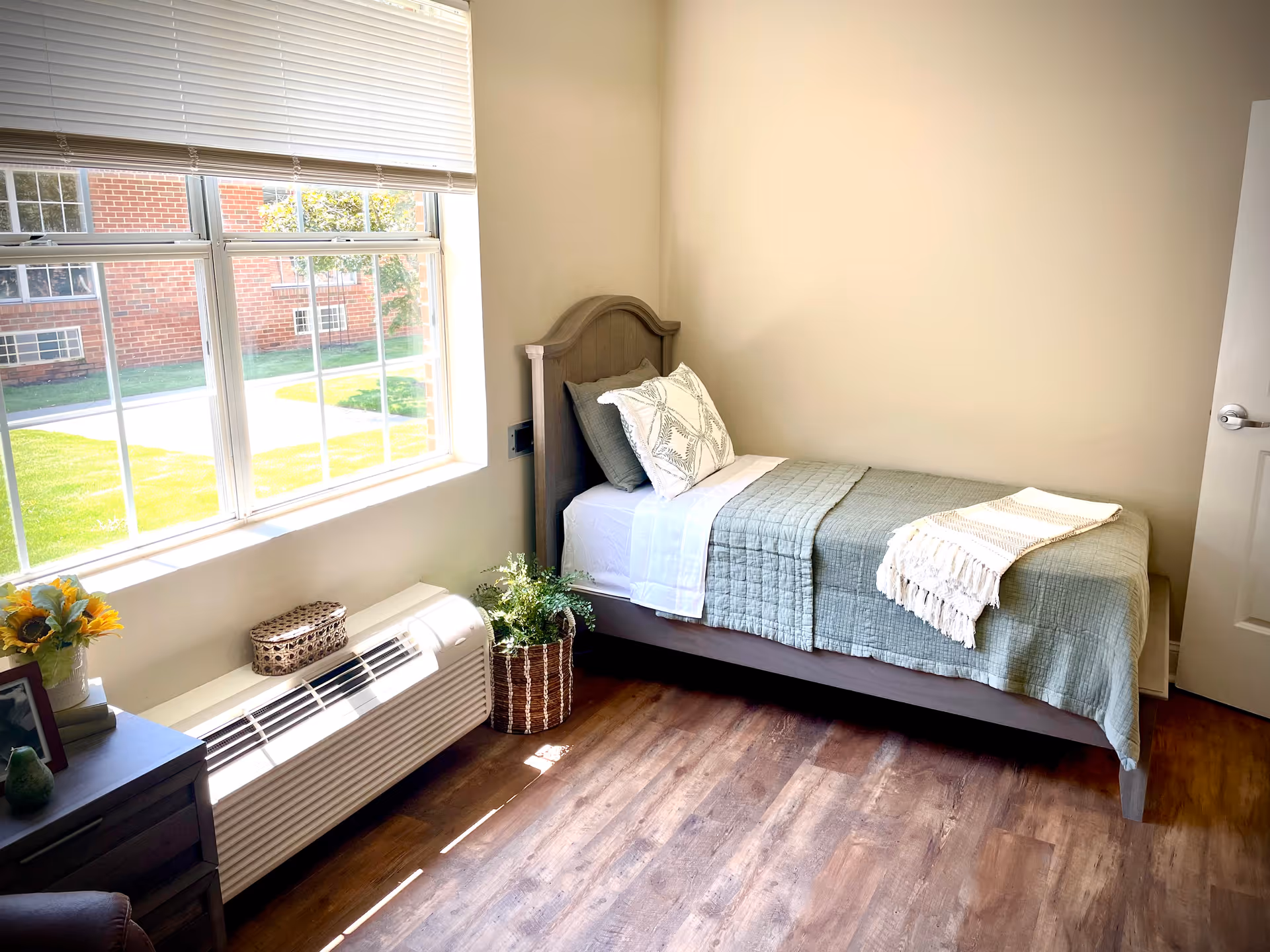 Sunlit small bedroom with a single twin bed against a beige wall, a large window, nightstand, and a potted plant.