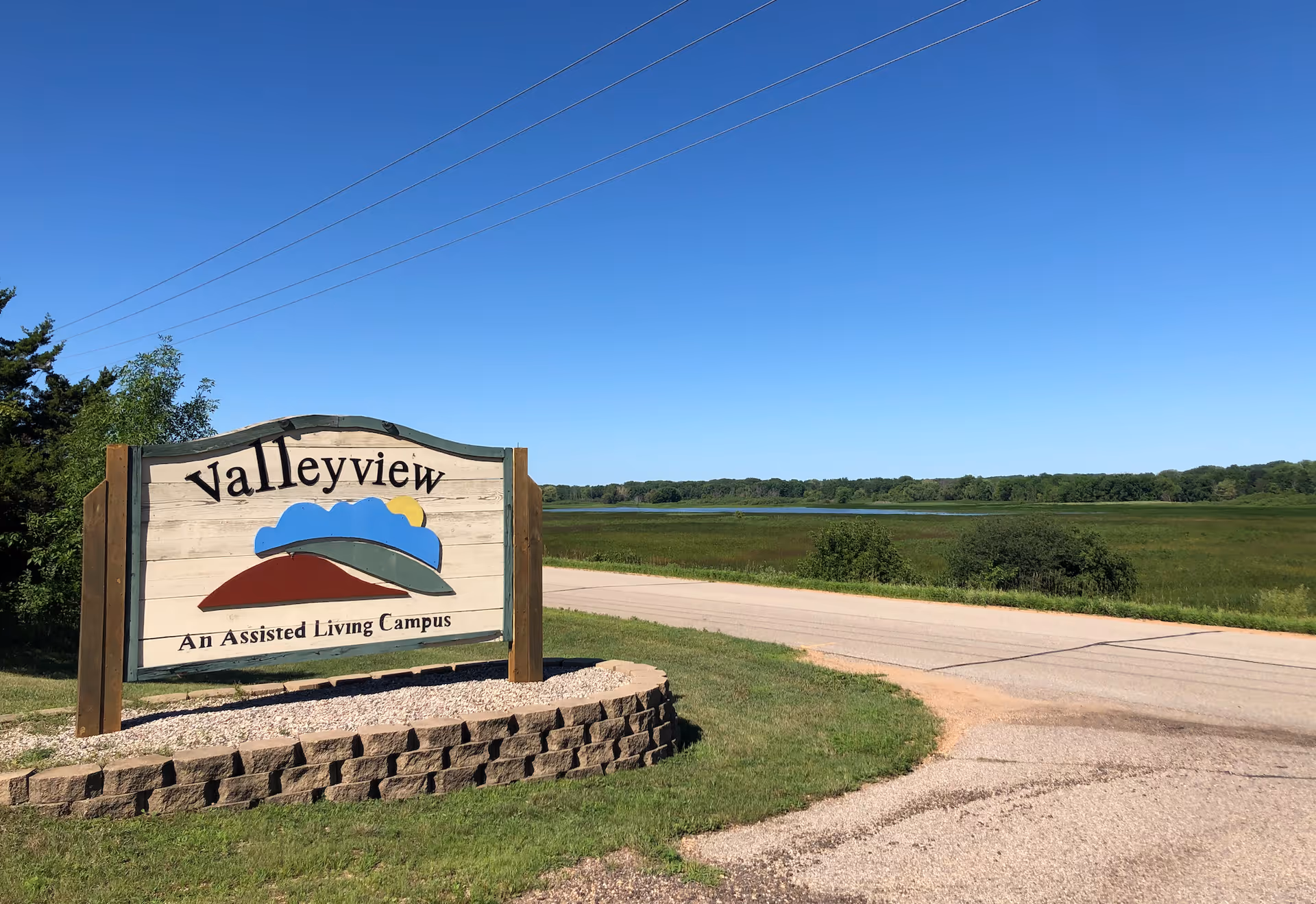 A wooden sign for Valleyview, an assisted living campus, stands beside a road with green grass and bushes around it. In the background, there is a wide open field with trees and a clear blue sky above.