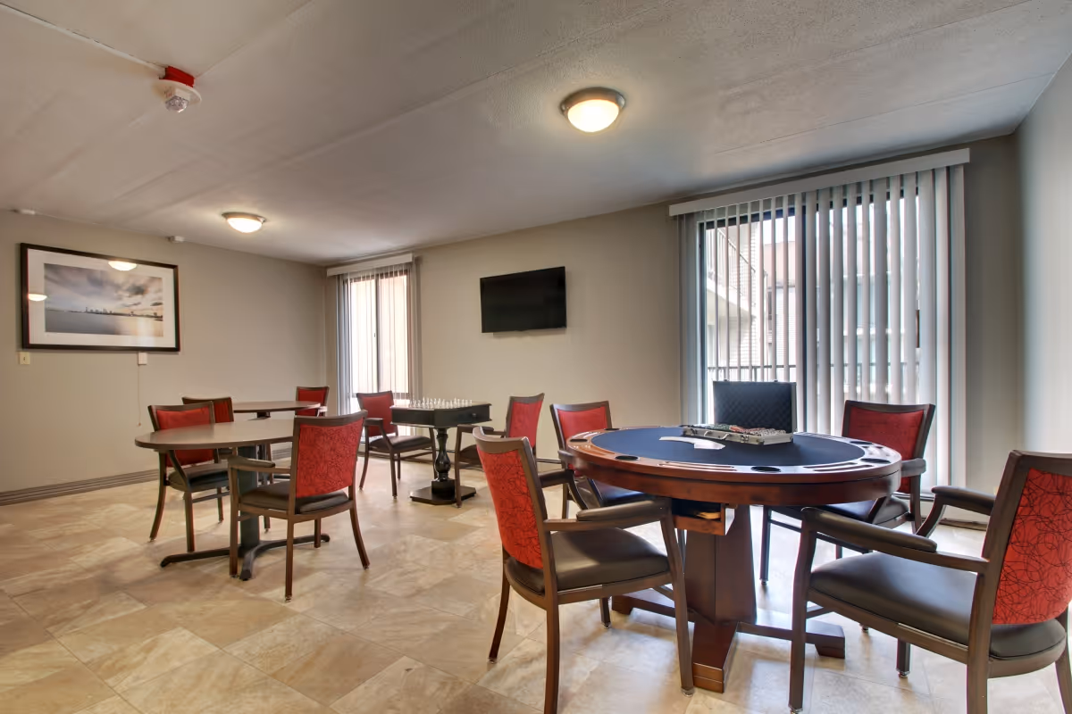 An interior activity room with multiple tables and red-upholstered chairs, a poker table in the foreground, a wall-mounted TV, and vertical blinds over sliding doors.
