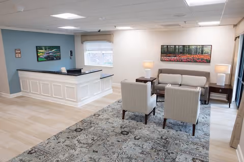 A bright and clean reception area in a senior living facility featuring a white reception desk with a black countertop, two beige armchairs, a beige sofa, two side tables with lamps, a large patterned rug, and framed landscape artwork on the walls.