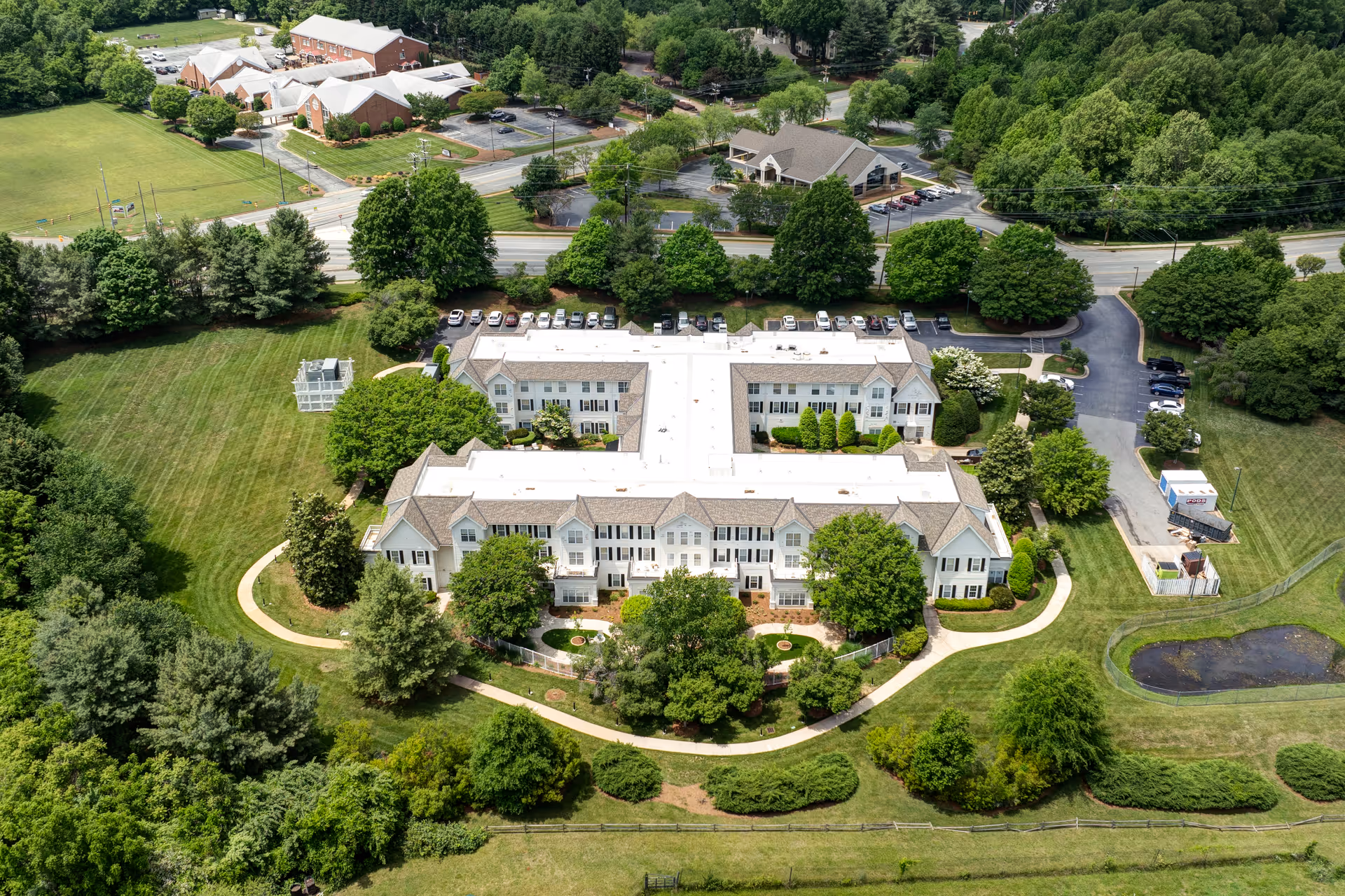 Aerial view of TerraBella Greensboro, a large senior living facility surrounded by green lawns, trees, and walking paths. The building is white with a light-colored roof and has multiple wings forming a U-shape. There are parking lots with cars and additional buildings visible in the background, along with a road and dense tree coverage around the property.
