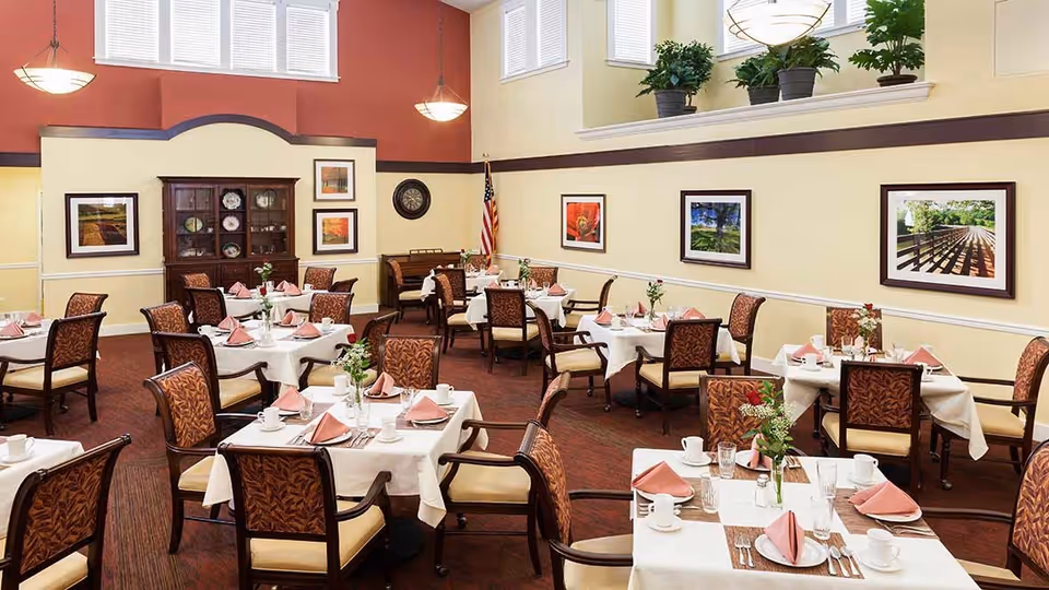 A dining room with multiple tables covered in white tablecloths, each set with pink folded napkins, plates, cups, and silverware. The room has high ceilings with large windows near the top, warm yellow and red walls, framed landscape pictures, and an American flag in the corner. There are cushioned chairs with patterned upholstery around each table, and a wooden cabinet with decorative plates is visible against the far wall.