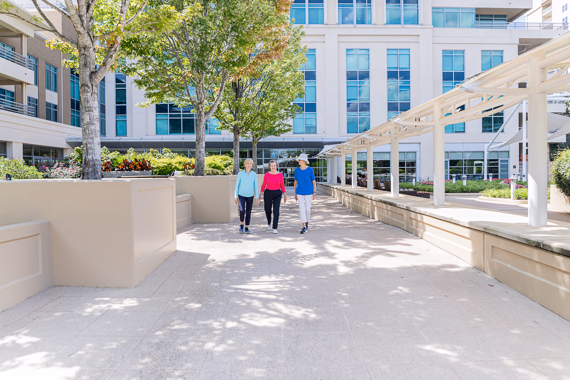Three people walking along a sunny courtyard beside a modern multi-story building.