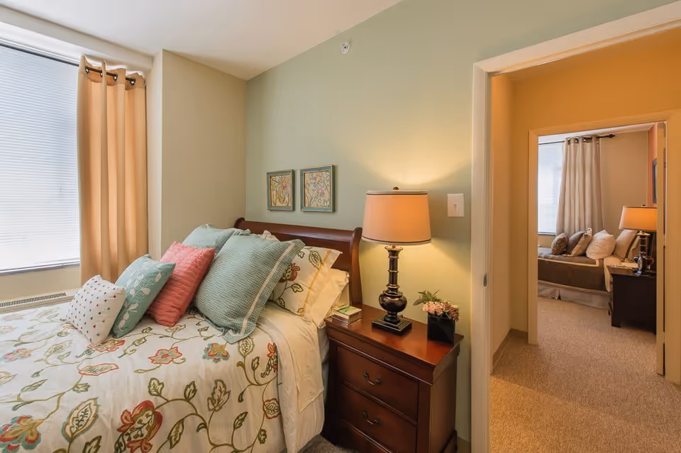 A cozy bedroom with a floral patterned bedspread and multiple pillows in pastel colors. There is a wooden nightstand with a lamp and a small plant next to the bed. The room has light green walls and a window with beige curtains. Through an open doorway, another bedroom with a similar setup is visible.