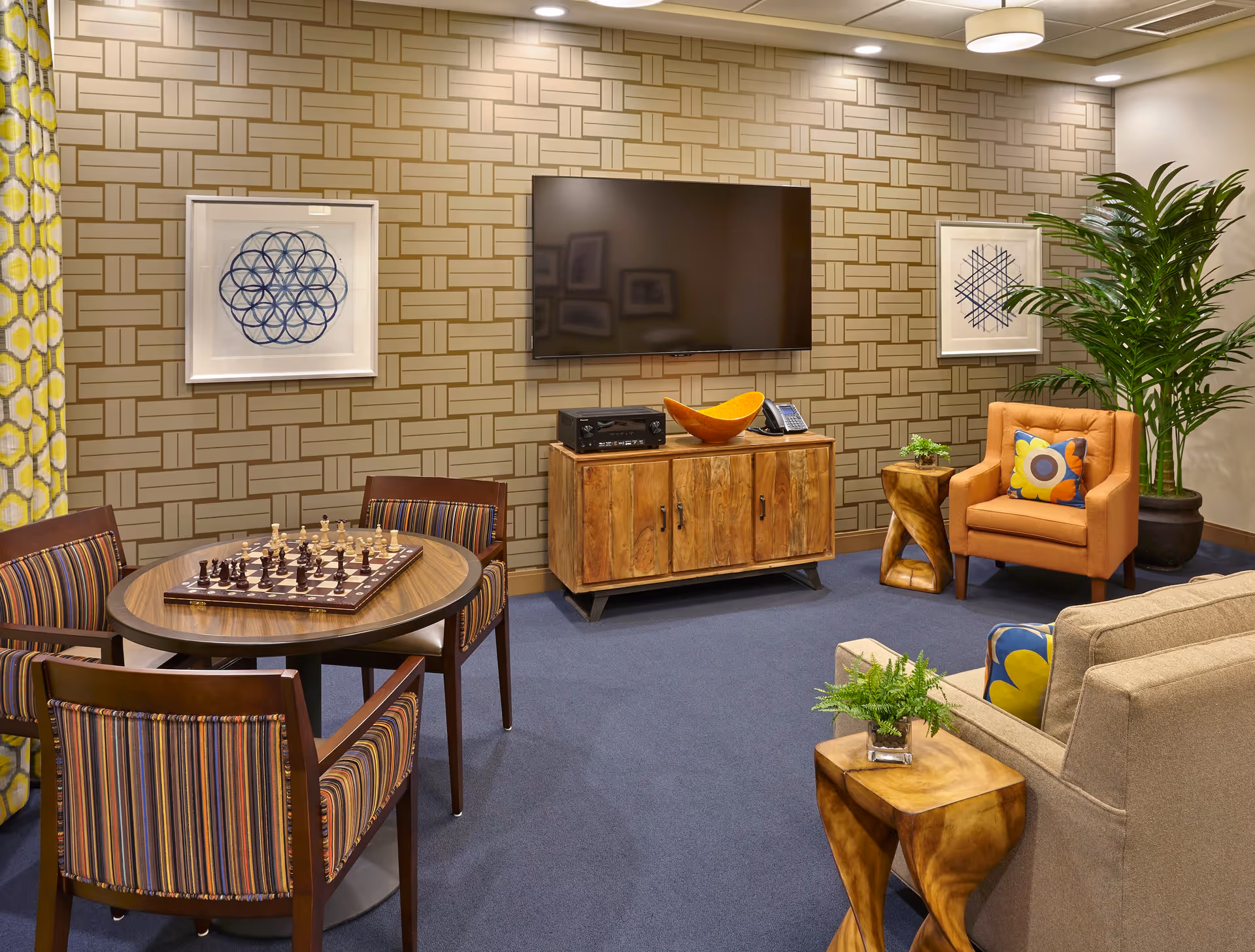 A cozy lounge with a wall-mounted TV, wooden media cabinet, round table with a chessboard and chairs, an armchair, sofa, and decorative plants.