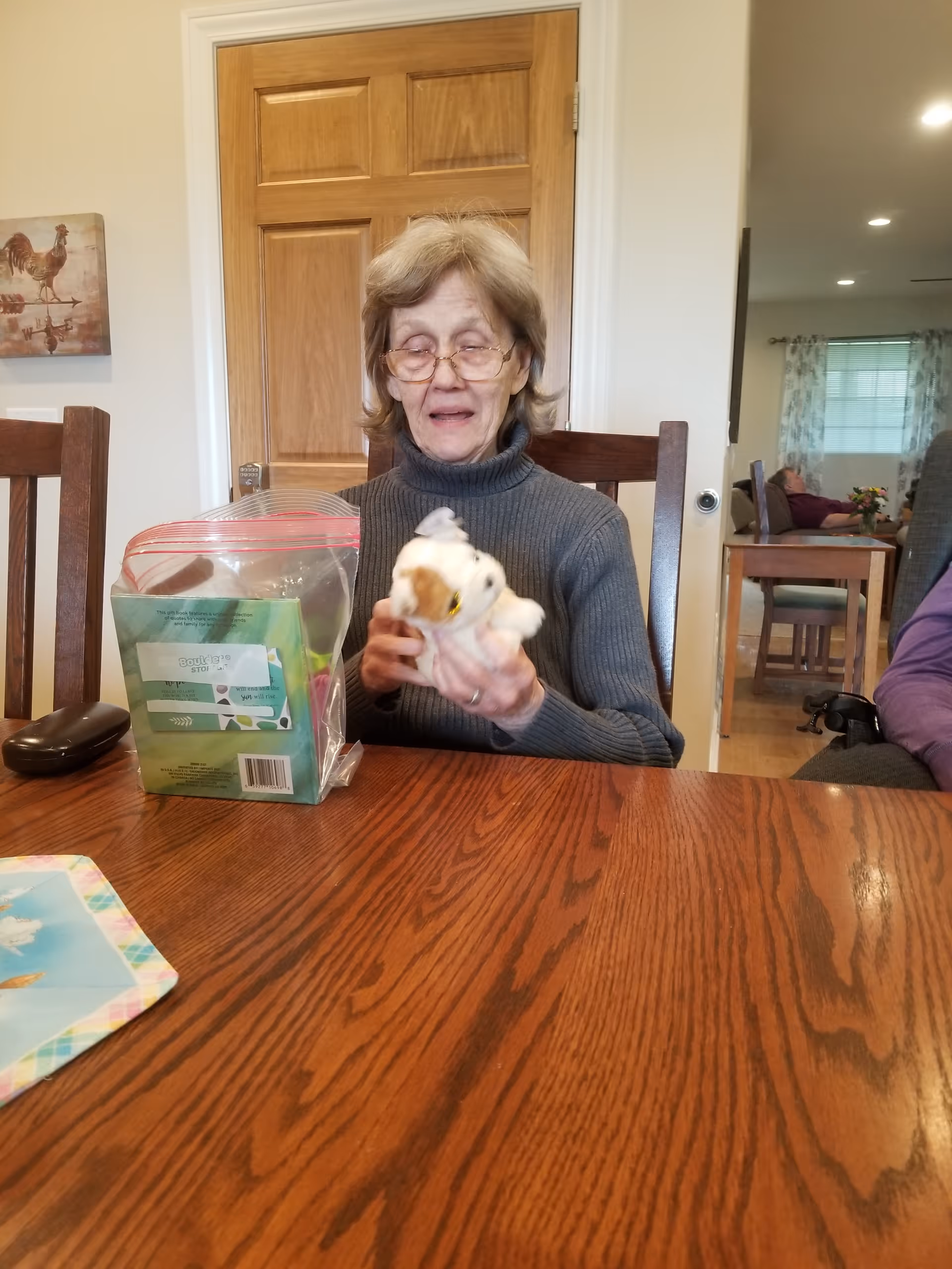 An elderly woman wearing glasses and a gray turtleneck sweater is sitting at a wooden table holding a small stuffed animal. On the table in front of her is a plastic bag with items inside and a remote control. Behind her is a wooden door and a wall with a painting of a rooster. In the background, another person is sitting on a chair near a window with curtains.