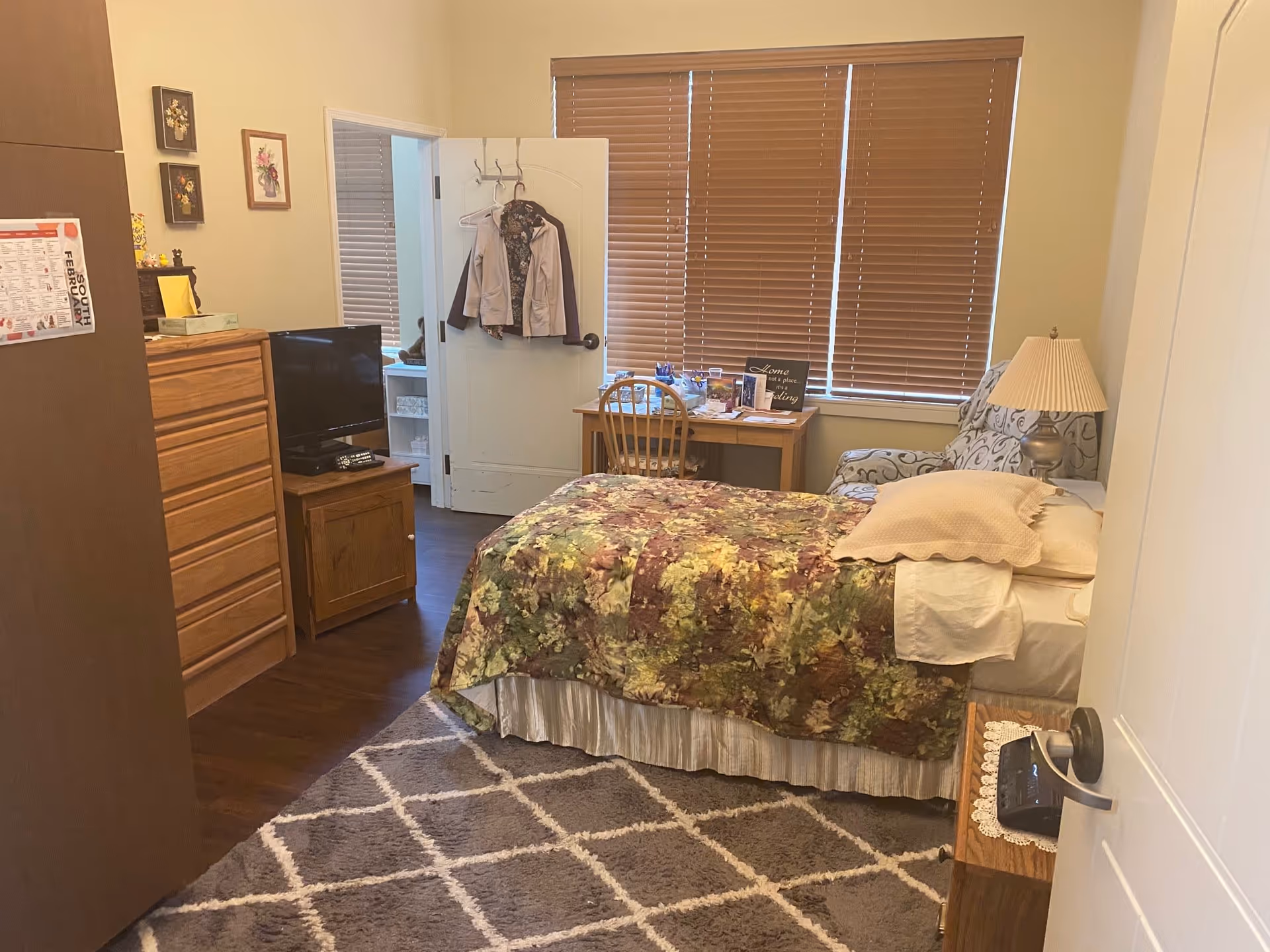 Cozy furnished bedroom with a bed, dresser, small TV, desk and armchair beneath closed blinds.