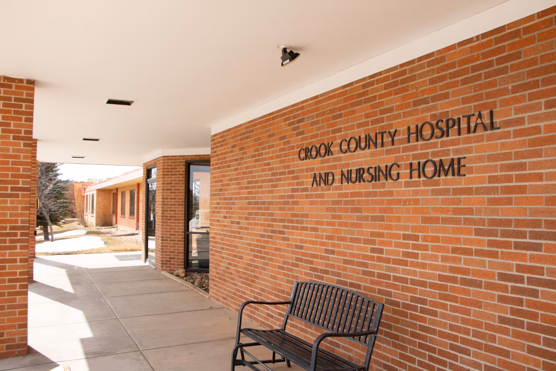 Covered entrance area of Crook County Hospital and Nursing Home with a brick wall displaying the facility name and a black metal bench placed against the wall.