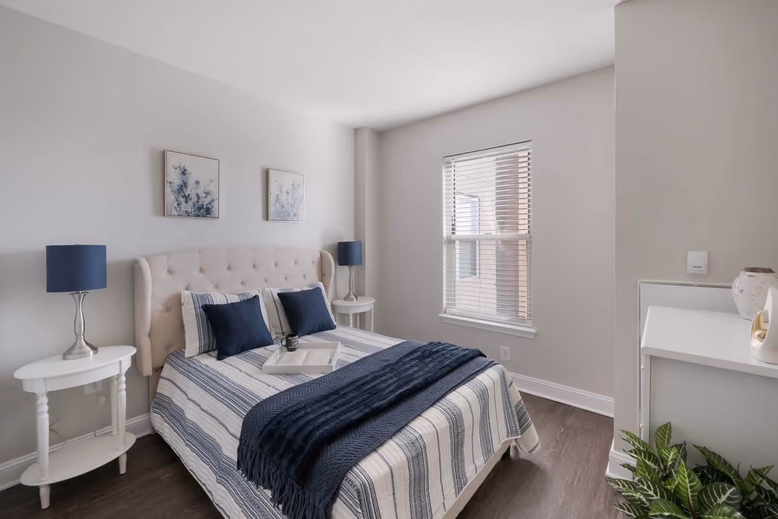 A neatly made bed with a tufted beige headboard, striped bedding in shades of blue and white, and two navy blue pillows. There are two white round bedside tables each with a blue lamp. Two framed floral artworks hang on the light gray wall above the bed. A window with white blinds is on the right wall, and a small white shelf with decorative items and a green plant is visible in the corner.