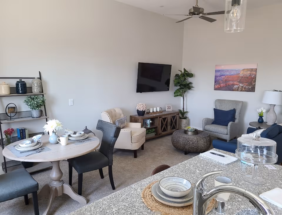 A cozy living room and dining area in a senior living facility. The room features a small round dining table set with plates, bowls, and mugs, surrounded by four chairs. In the living area, there are two armchairs, a blue sofa, a round woven coffee table, a TV mounted on the wall, and a wooden console table with decorative items. A tall green plant and a colorful landscape painting adorn the walls. The foreground shows a kitchen counter with a sink, dishes, and a glass cake stand.