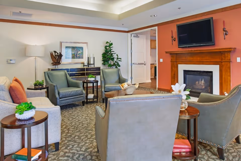 A cozy living room area with a beige sofa and green armchairs arranged around a wooden coffee table. The room features a fireplace with a wooden mantel and a flat-screen TV mounted above it. There are side tables with decorative plants and books, a floor lamp, a framed artwork on the wall, and a green potted plant near a doorway leading to another room.