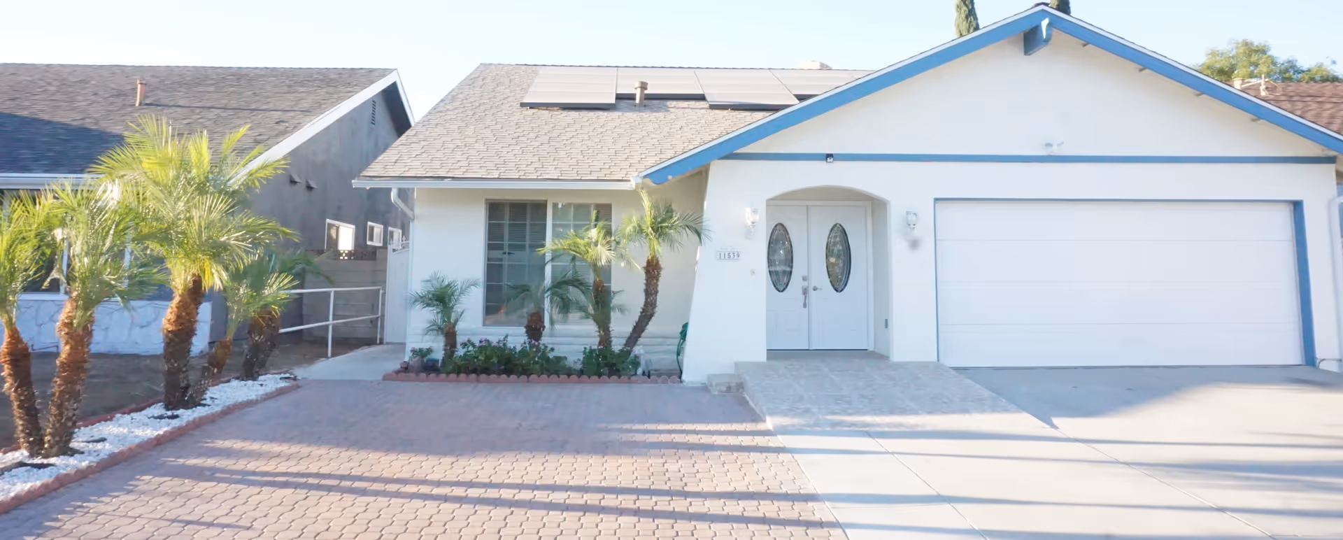 Front exterior view of a single-story residential building with a two-car garage, white walls, blue trim, solar panels on the roof, a paved driveway, and small palm trees and plants in the front yard.