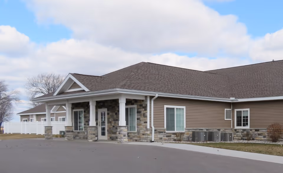 Single-story assisted living building entrance with a covered porch, stone veneer columns, and brown siding under a cloudy sky.