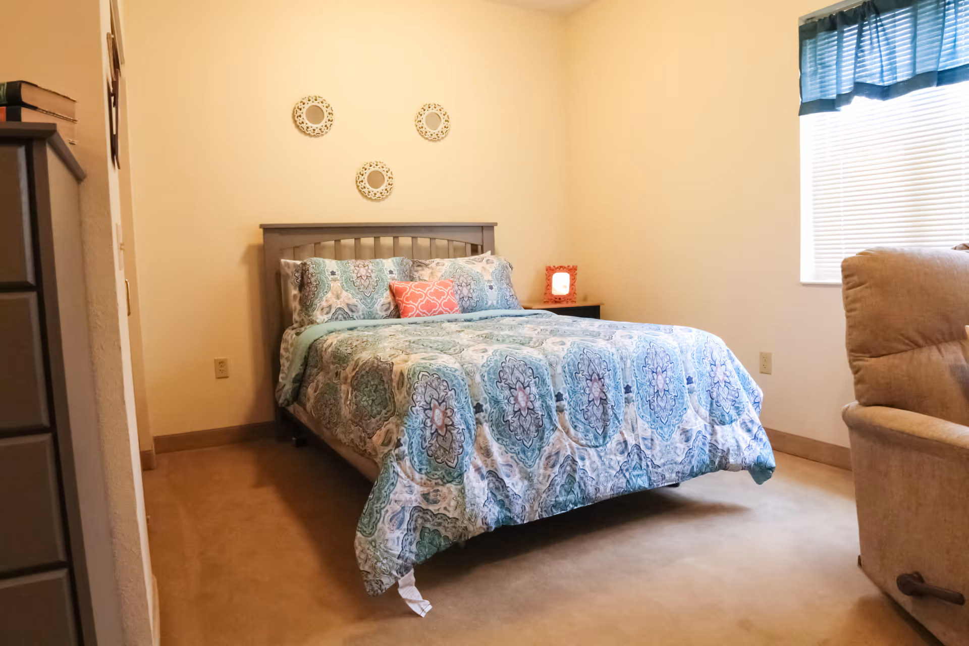 A bedroom with a bed covered in a blue and green patterned comforter and pillows, a small nightstand with a red-framed clock, three decorative round mirrors on the wall above the bed, a window with blinds and a blue valance, a beige recliner chair, and a tall dresser partially visible on the left.