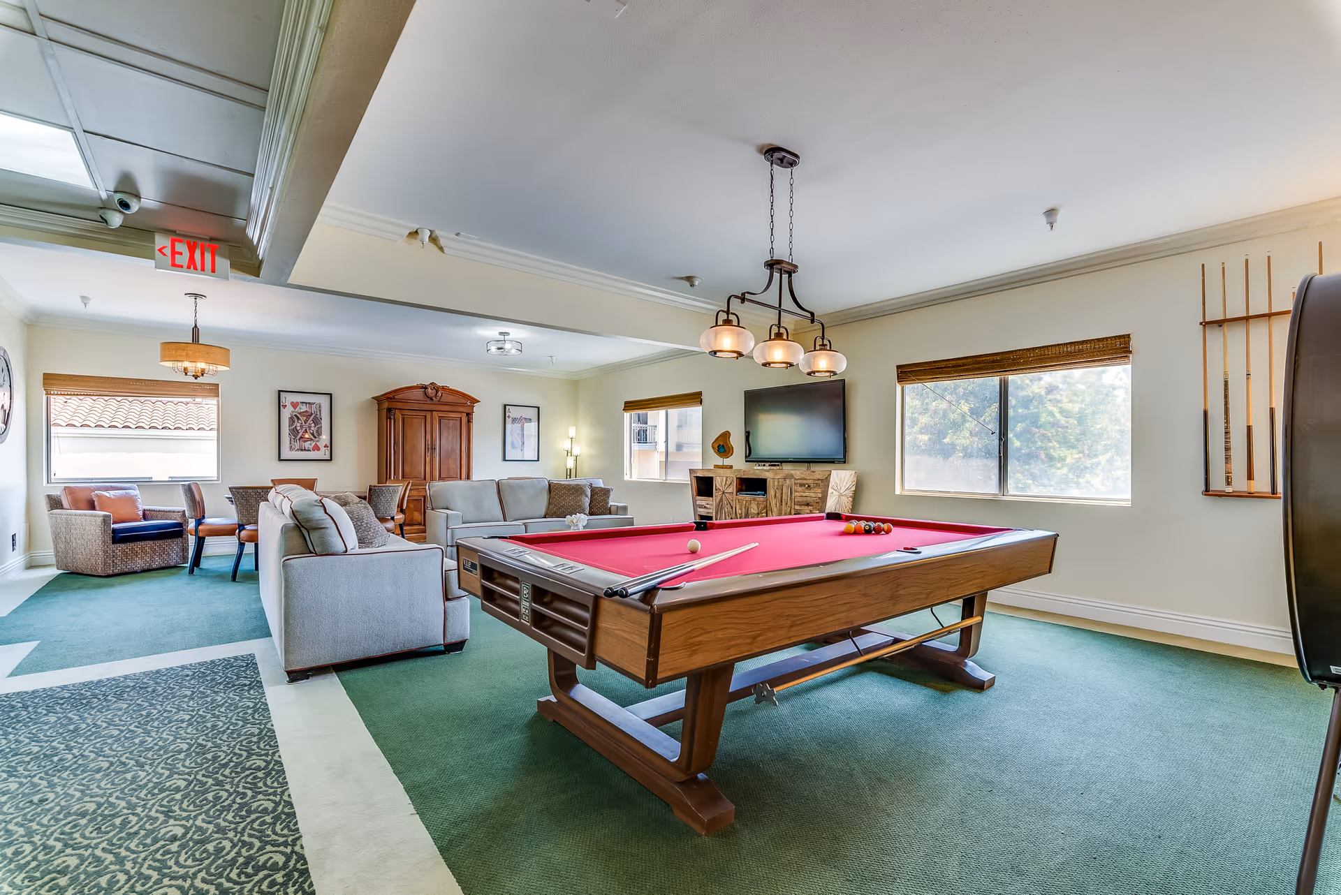 A bright and spacious senior living common area featuring a pool table with a red felt surface in the foreground. Behind the pool table, there are two beige sofas arranged around a wooden cabinet with a flat-screen TV mounted above it. The room has large windows with natural light coming in, green carpet flooring, and a ceiling light fixture above the pool table. On the right wall, there is a rack holding pool cues. In the background, there is a small seating area with chairs and a table near an exit sign.