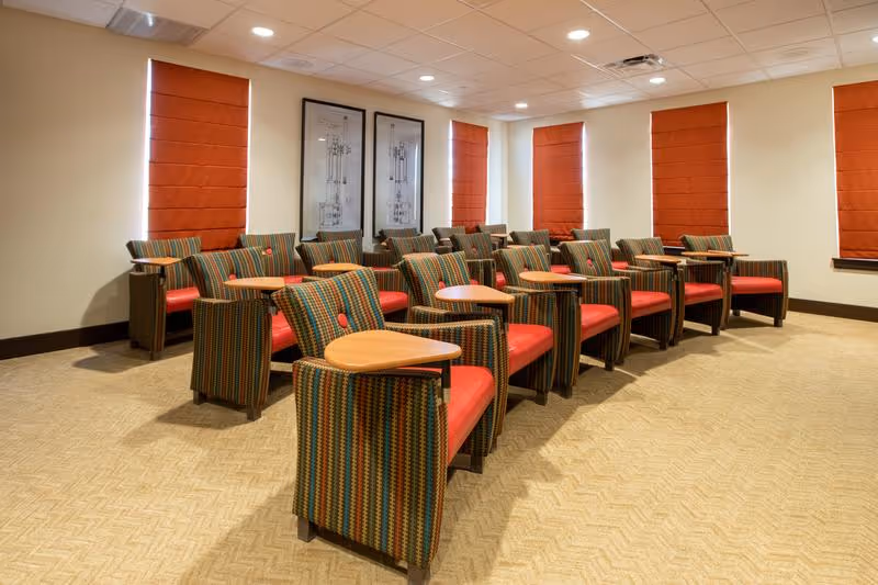 A room with multiple rows of striped armchairs featuring red cushions and attached wooden writing surfaces, arranged facing the same direction. The room has beige carpet, cream-colored walls, red window shades, and two framed architectural drawings on the wall.