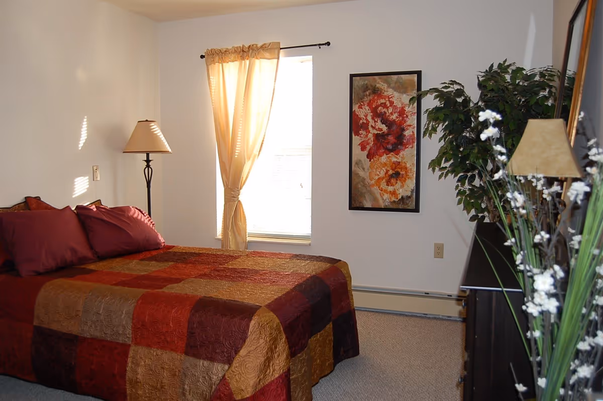 A bedroom with a bed covered in a patchwork quilt in shades of red, brown, and gold. There are two matching pillows on the bed. A floor lamp stands next to the bed near a window with beige curtains. A framed floral painting hangs on the wall, and a large green plant is placed near a dresser with a lamp and decorative white flowers.