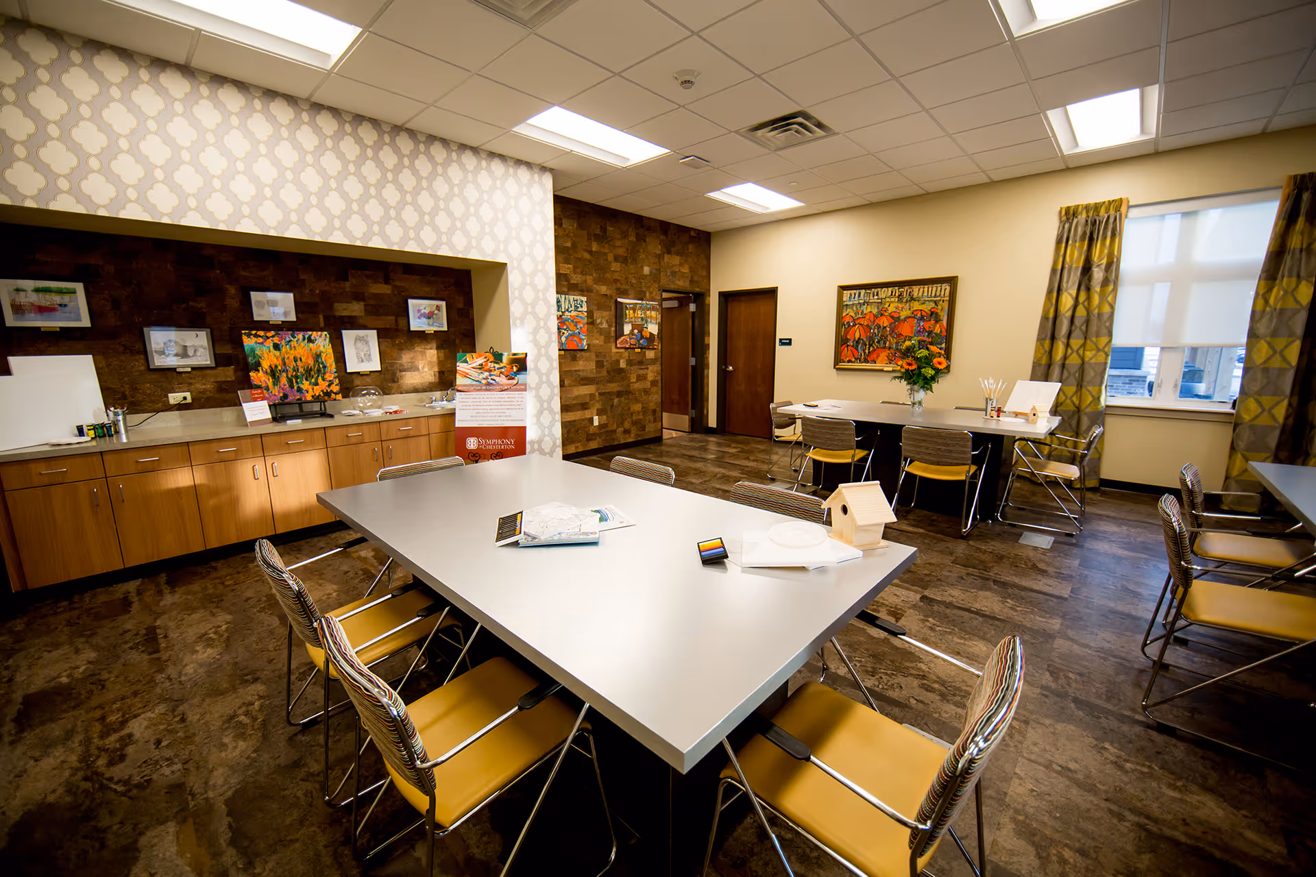 Interior room with modern decor featuring two large tables surrounded by chairs with yellow seats. The room has wooden cabinets along one wall with various artworks displayed above them. There are patterned curtains on the windows and framed paintings on the walls. The floor has a dark wood finish and the ceiling has recessed lighting.