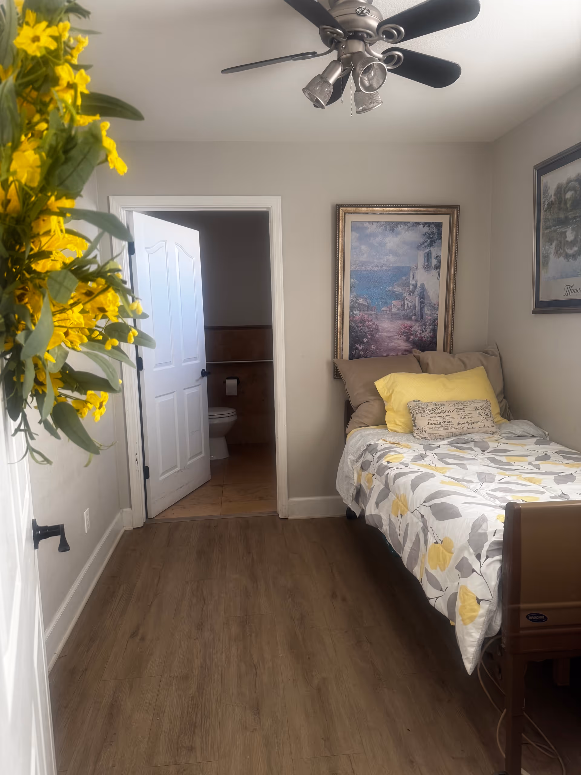 A small bedroom with a single bed covered in a floral-patterned comforter with yellow and gray colors. There are two pillows on the bed, one yellow and one with text. The room has light-colored walls with two framed paintings hanging. A ceiling fan with lights is mounted on the ceiling. The door to an adjoining bathroom is open, showing a toilet and tiled floor. A bunch of yellow flowers is visible on the left side of the image.