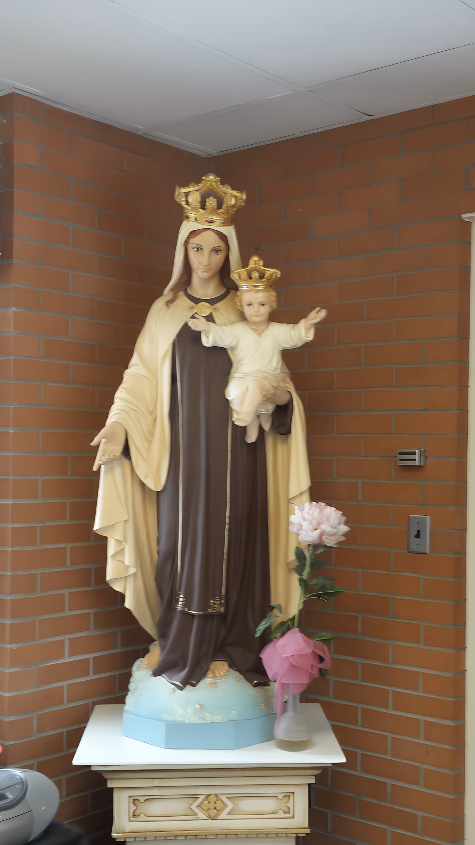 A statue of the Virgin Mary holding baby Jesus, both wearing golden crowns, standing on a pedestal in front of a brick wall. There is a pink flower in a vase with a pink ribbon placed on the pedestal next to the statue.