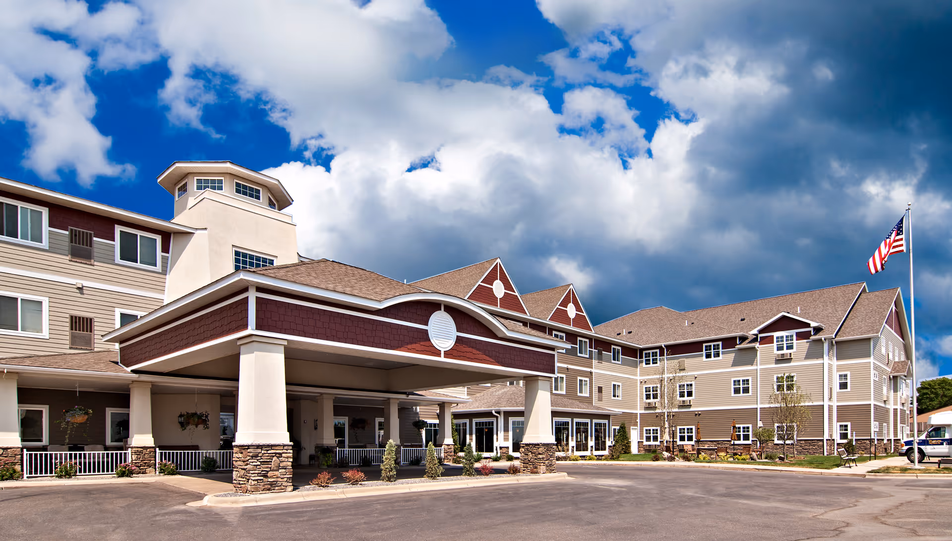 Exterior view of a large senior living facility building with multiple windows, a covered entrance supported by columns with stone bases, and an American flag on a flagpole. The sky is partly cloudy with blue patches.
