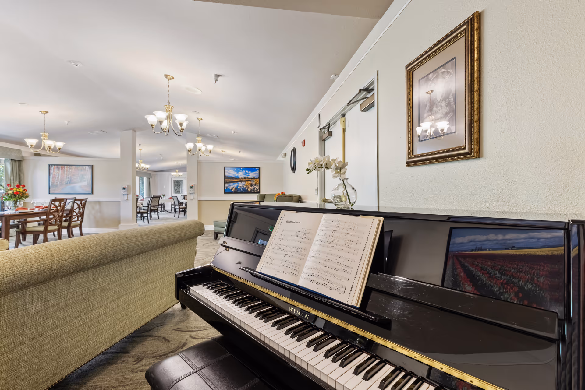 Interior view of a senior living facility common area featuring a black Wyman piano with sheet music on the stand, a beige couch, dining tables with chairs, chandeliers hanging from the ceiling, and framed artwork on the walls.