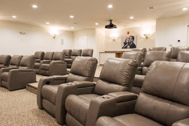 A cozy theater room with multiple rows of plush, brown reclining chairs arranged in tiers facing a projector. The room has beige walls, carpeted floors, and soft wall sconces providing warm lighting. A black and white framed picture is visible on the back wall.