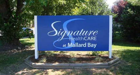 Blue sign reads Signature HealthCARE at Mallard Bay on a grassy outdoor display.