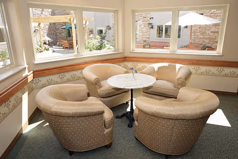 A cozy seating area with four beige upholstered armchairs arranged around a small round white marble table. The space is well-lit by natural light coming through large windows that offer a view of an outdoor patio with benches, umbrellas, and greenery.