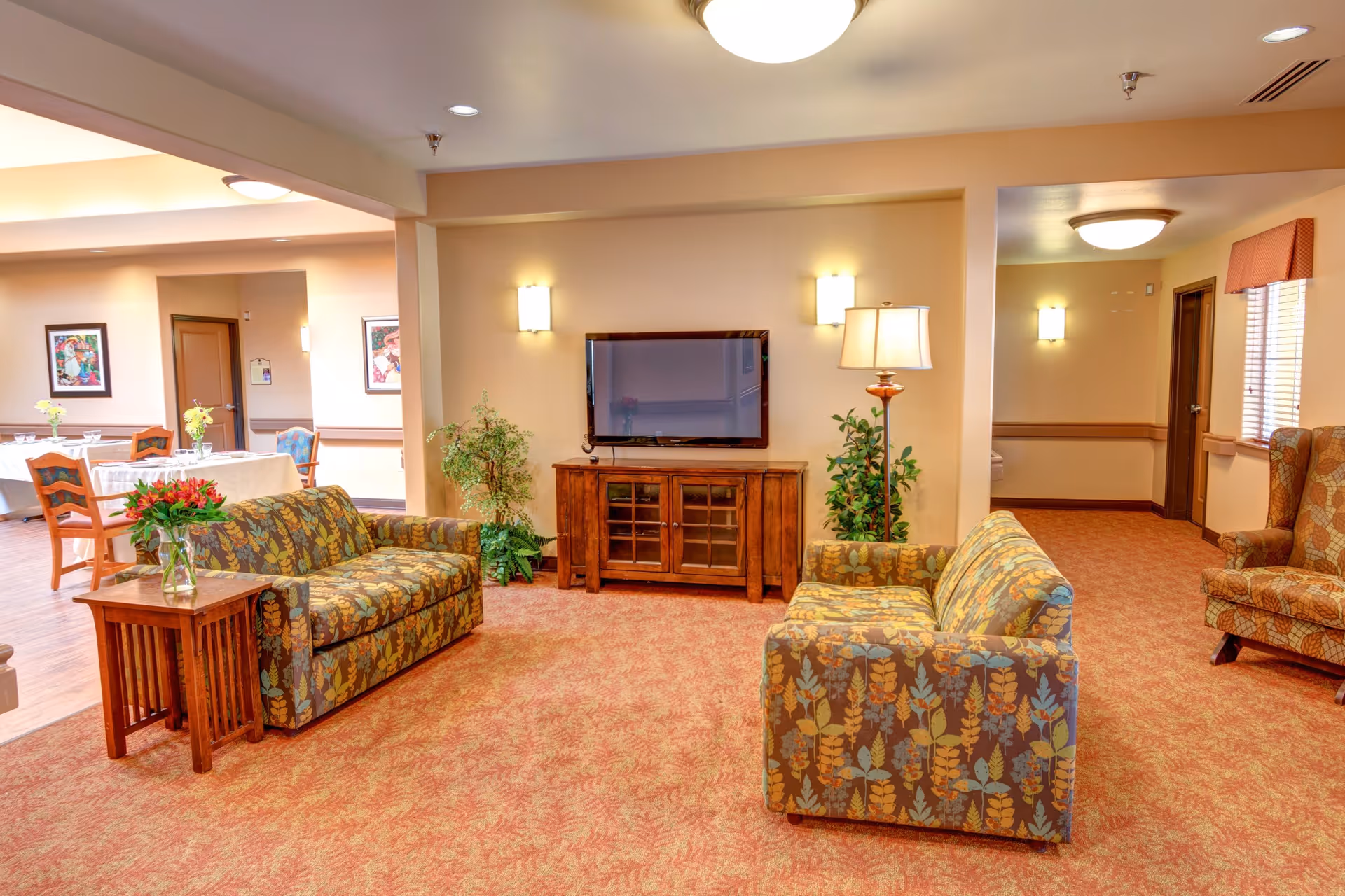 A cozy living room area in a senior living facility with two floral-patterned sofas facing a wall-mounted flat-screen TV above a wooden cabinet. There is a wooden side table with a vase of flowers, a floor lamp, potted plants, and warm lighting. In the background, a dining area with tables and chairs is visible.