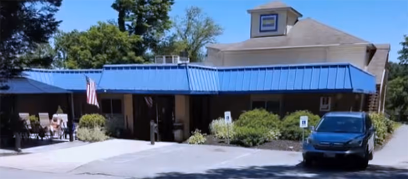 Exterior view of a single-story building with a blue metal roof and an American flag displayed near the entrance. There is a parking area in front with a black car parked and some greenery around the building.