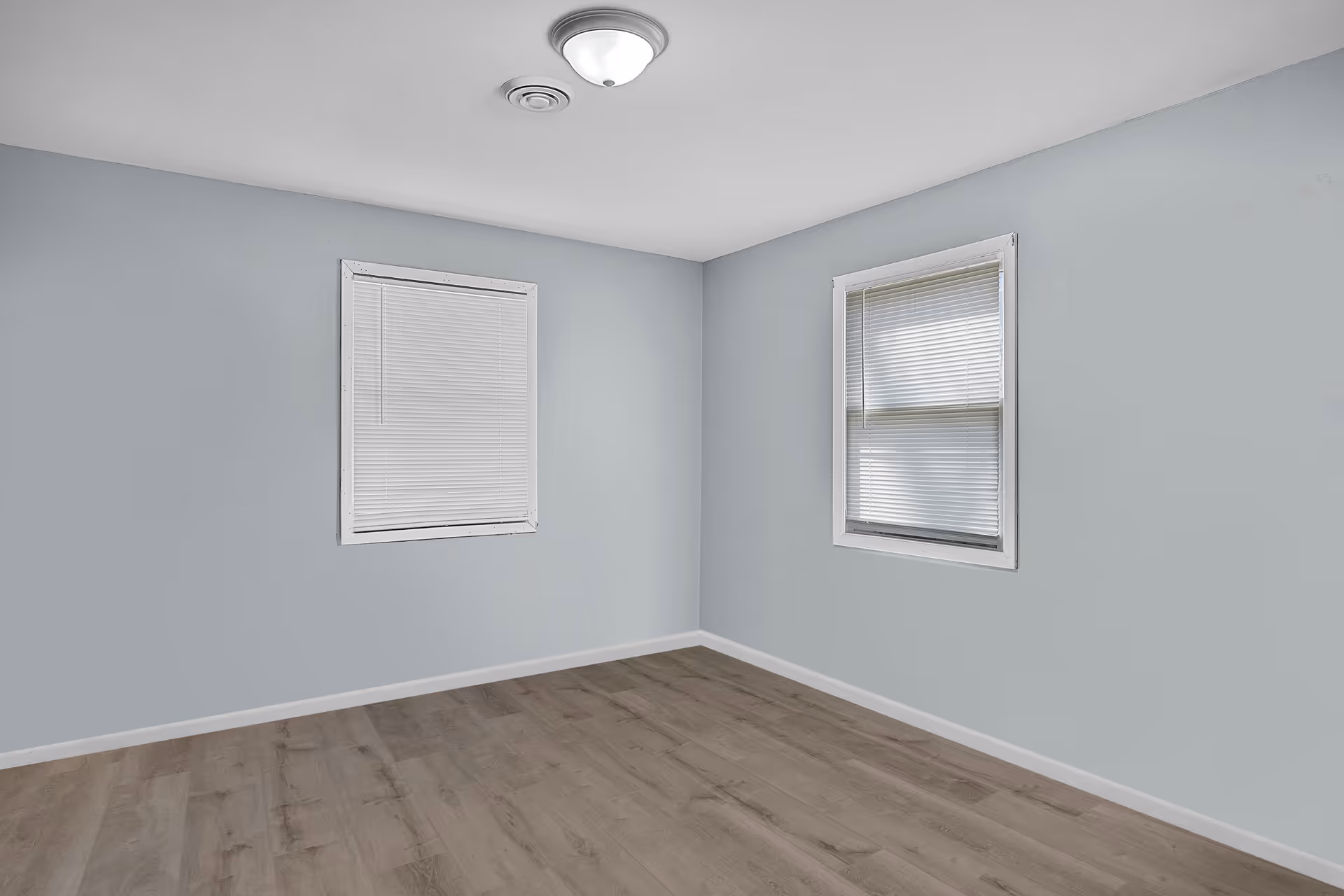 Empty room with light gray walls, two windows with white blinds, a ceiling light fixture, and wood-look flooring.