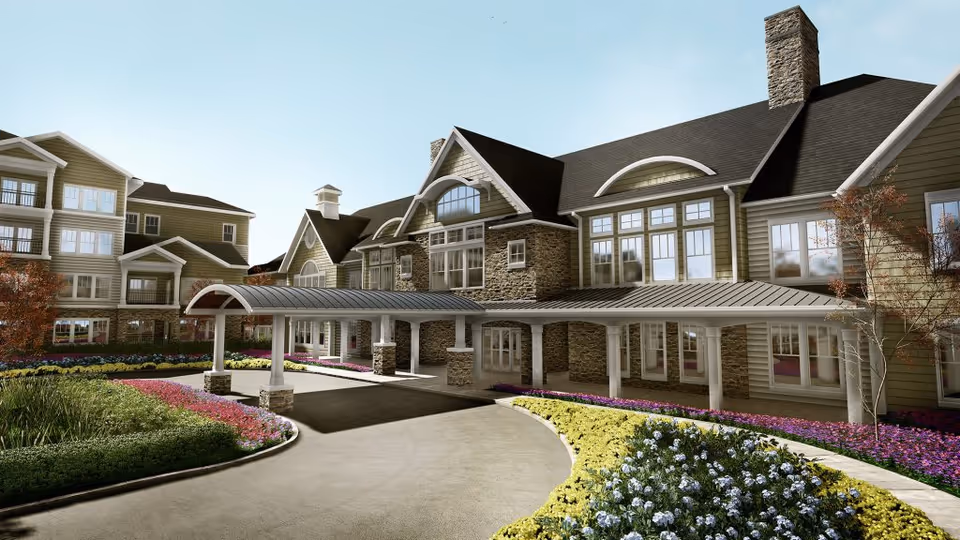 Exterior view of a senior living facility building with a covered entrance driveway, stone and wood siding, multiple windows, and landscaped flower beds with colorful flowers and greenery under a clear sky.
