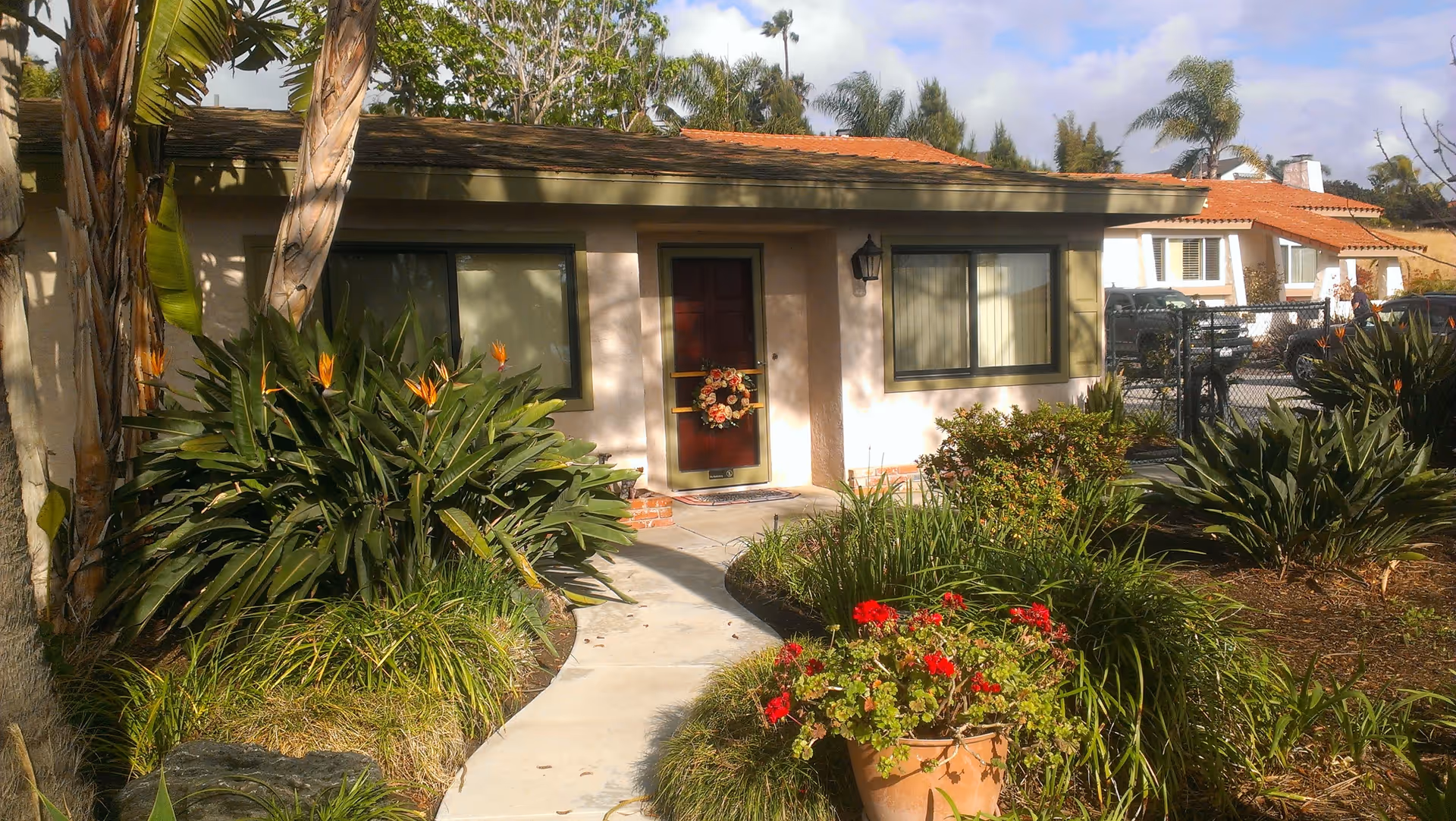 A single-story building with a brown door decorated with a floral wreath, two windows with closed blinds, and a pathway leading to the entrance. The front yard features lush green plants, including bird of paradise flowers and a potted plant with red flowers. In the background, there are palm trees, other houses, and parked cars.
