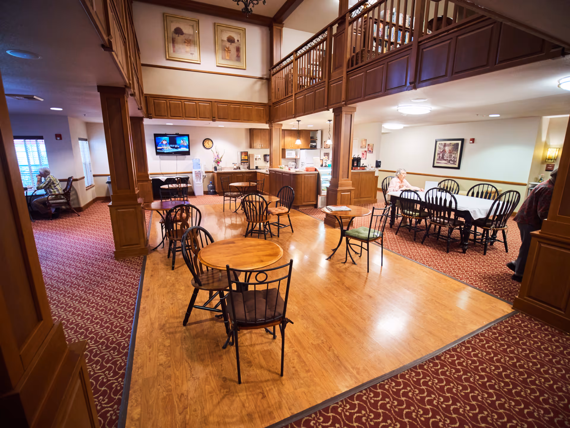 A common area in a senior living facility with wooden tables and chairs arranged on a wooden floor section surrounded by carpet. There is a kitchenette area with cabinets, a microwave, coffee machine, and a water cooler. Two elderly women are seated at tables, one on the left near windows and another on the right side. The room has warm lighting, framed pictures on the walls, and a television mounted on the wall.