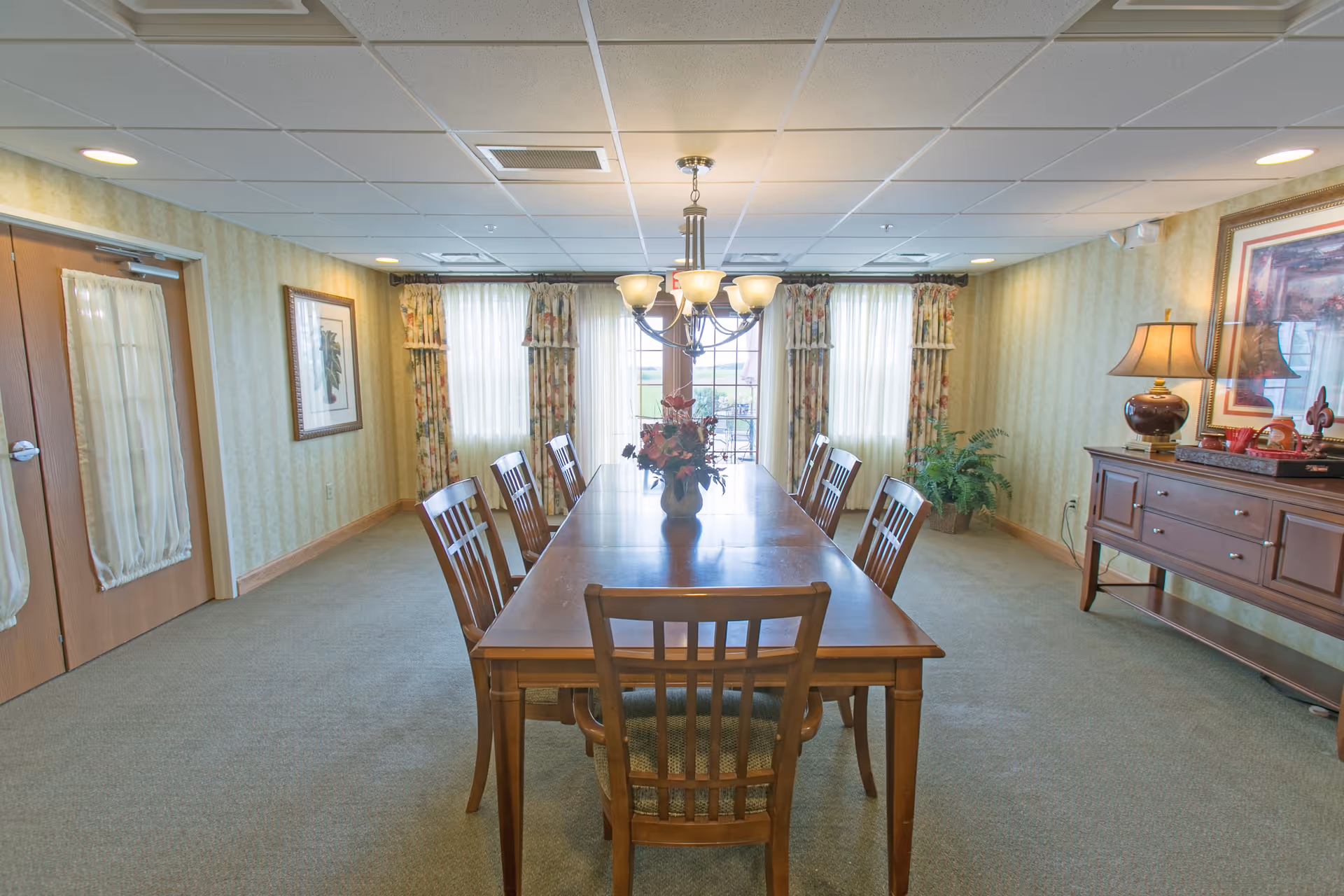 A dining room with a long wooden table surrounded by eight matching wooden chairs. A chandelier with multiple lights hangs above the table, which has a floral centerpiece. The room has carpeted floors, patterned wallpaper, and large windows with floral curtains allowing natural light to enter. On the right side, there is a wooden sideboard with a lamp, framed artwork, and decorative items. Double doors with sheer curtains are on the left wall.