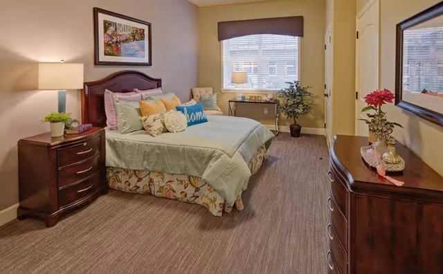 A warmly decorated bedroom with a made bed, bedside tables, a dresser, lamp, and a window with blinds.