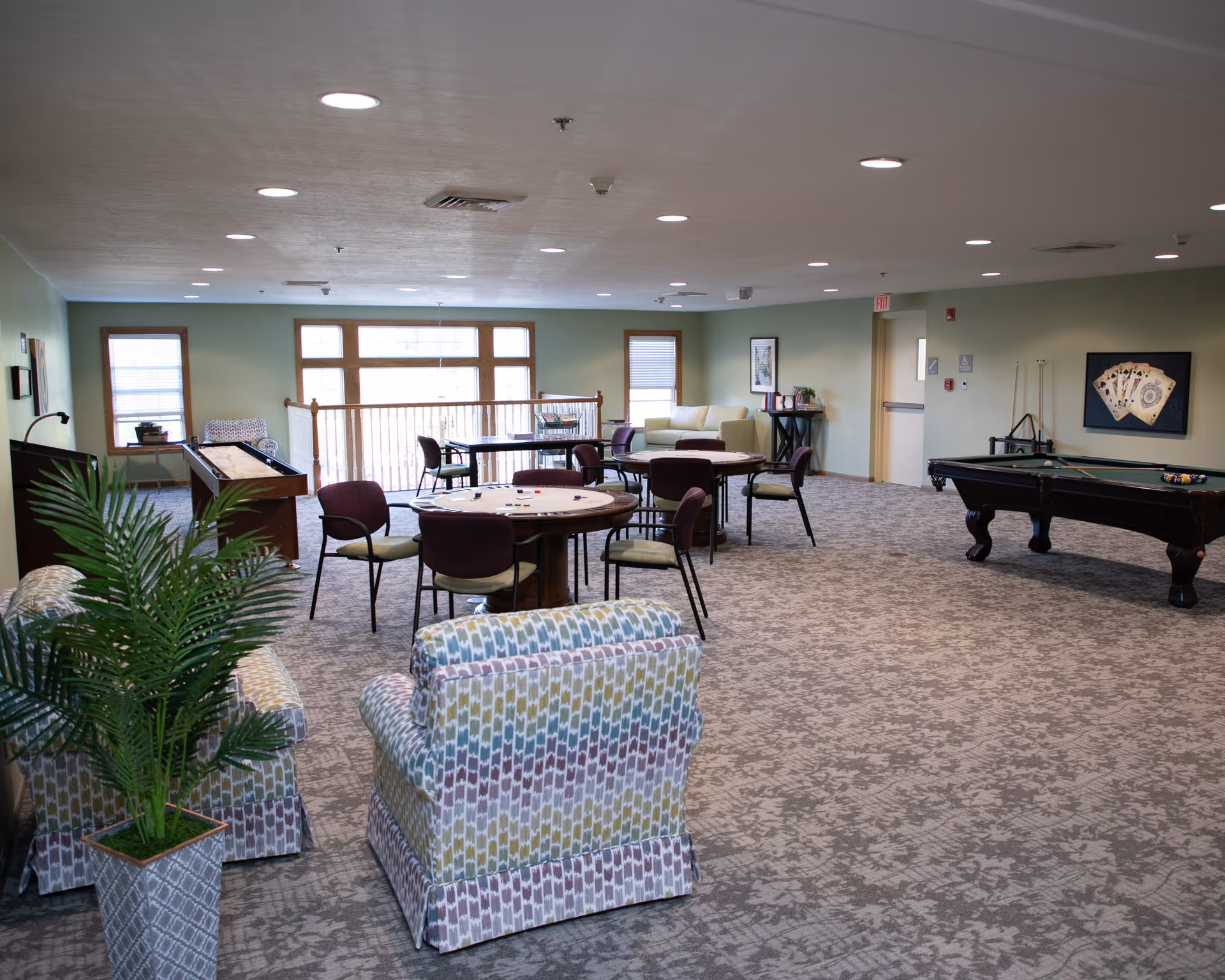 A spacious recreational room with patterned armchairs, round tables with chairs, a shuffleboard table, and a pool table. The room has large windows letting in natural light, green walls, and carpeted floors. There is also a framed picture of playing cards on the wall.