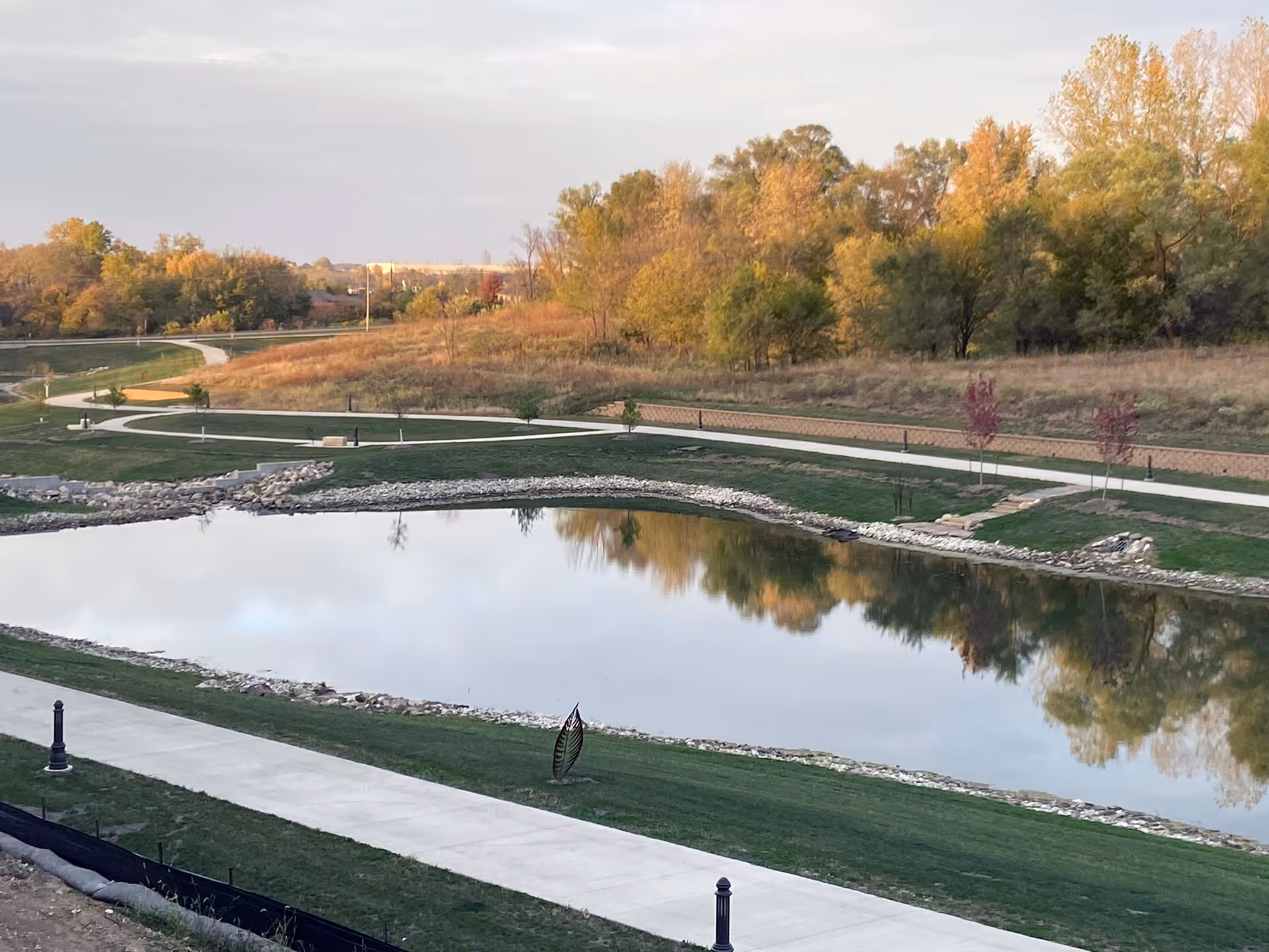 A serene outdoor scene at Holland Farms Senior Living featuring a calm pond surrounded by green grass and walking paths. Trees with autumn foliage are visible in the background under a partly cloudy sky.