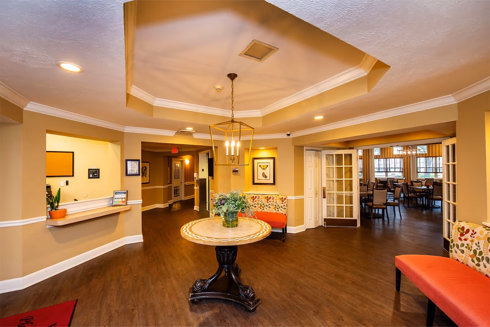 Interior view of a senior living facility lobby area with a round table holding a floral arrangement in the center. The space features warm beige walls, wood flooring, recessed lighting, and a decorative ceiling light fixture. There are cushioned benches with floral upholstery along the walls and a reception window on the left. In the background, a dining area with tables and chairs is visible through open double doors.