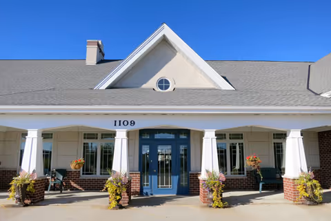 Front entrance of a single-story senior living facility with blue double doors, white columns, brick accents, planters, and the address number 1109 under a peaked roof.