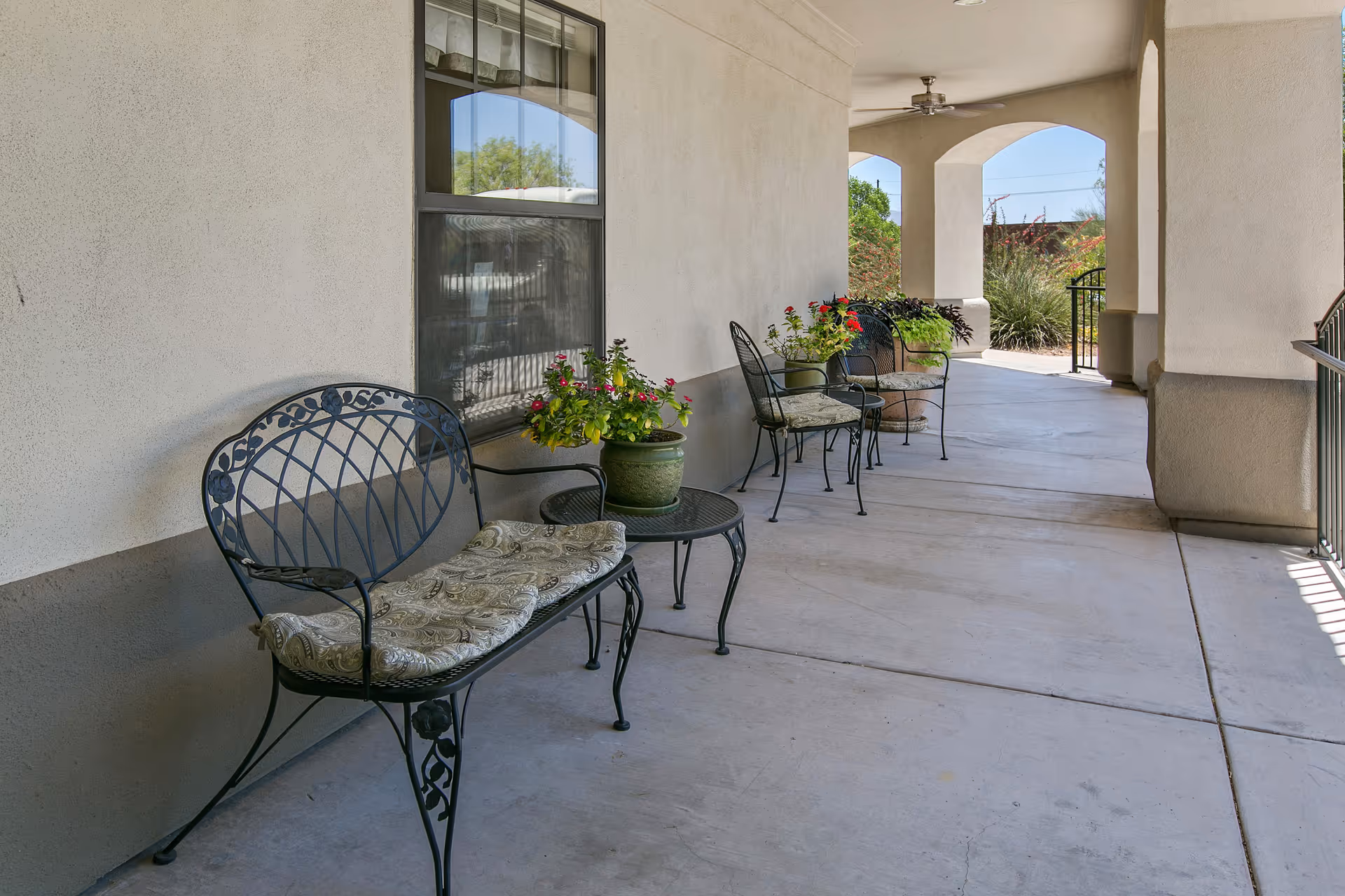 Covered outdoor patio area with metal benches and chairs featuring cushions, small tables with potted plants, and a ceiling fan. The patio has a concrete floor and is adjacent to a building with beige walls and windows.