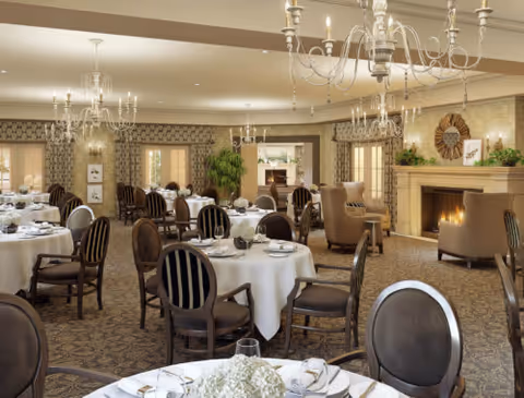 Elegant dining room with round tables covered in white tablecloths, set with plates, glasses, and silverware. The room features upholstered chairs with dark wood frames, chandeliers hanging from the ceiling, patterned carpet, and a cozy fireplace with decorative items on the mantel. Large windows and doorways with curtains allow natural light into the space.