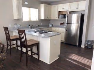 A modern kitchen area with white cabinets, stainless steel refrigerator and microwave, a kitchen island with a cooktop, and two wooden chairs with cushions. The floor is dark wood, and there are windows with blinds letting in natural light.