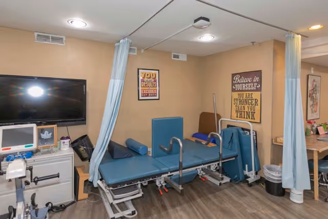 A therapy or rehabilitation room with a padded treatment table equipped with support cushions and handrails. The room has beige walls with motivational posters, a large flat-screen TV mounted on the wall, medical equipment on a cabinet, and light blue privacy curtains. The floor is wood-patterned.